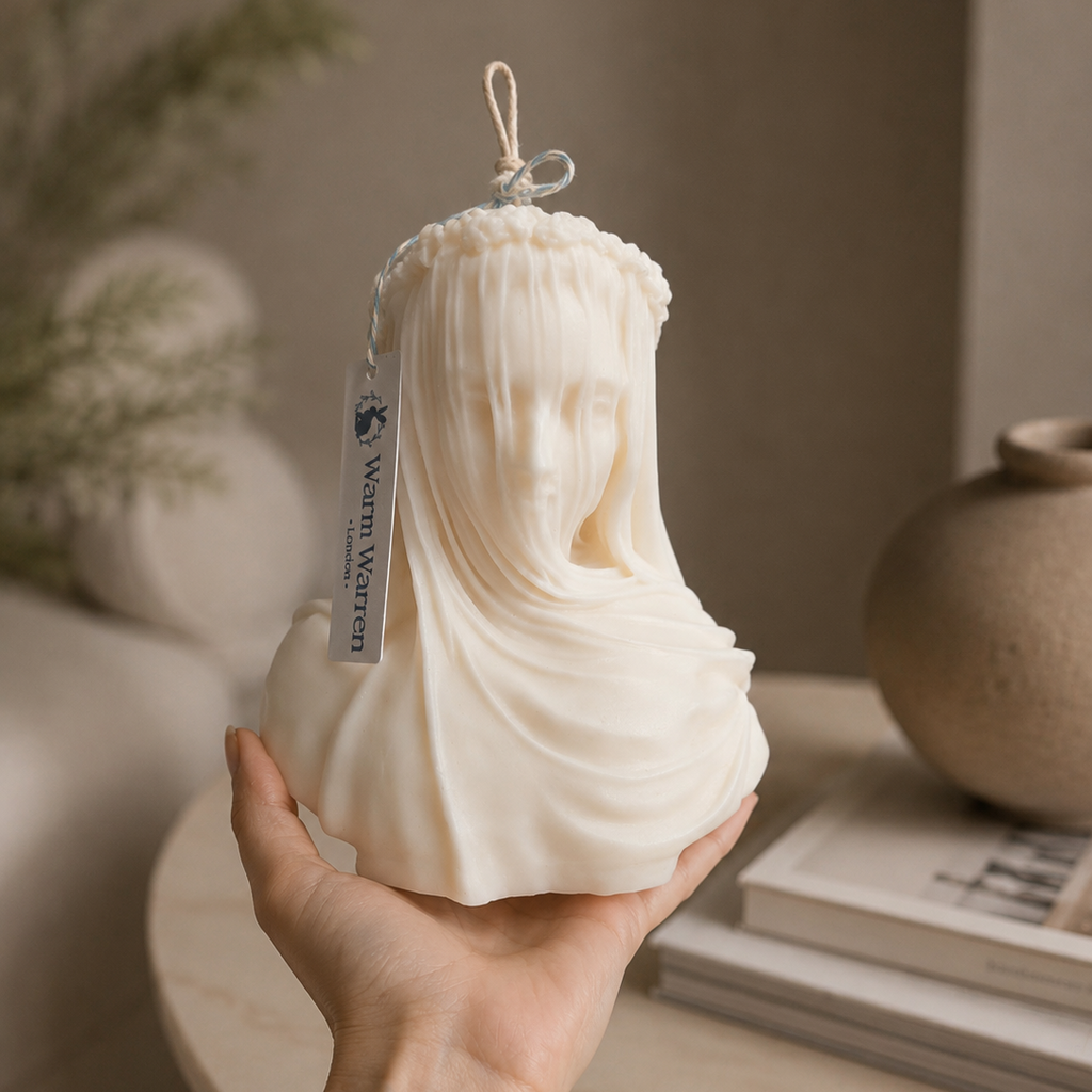 A hand holds a cream-colored wax candle shaped like a classical bust of a draped female figure. Warm Warren London Veiled Vestal soy wax candle