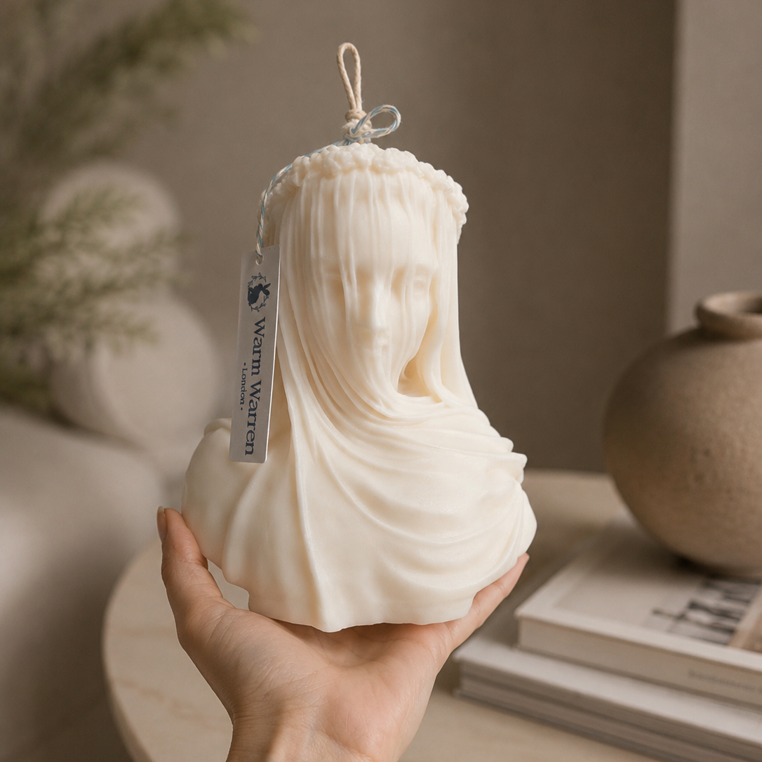 A hand holds a cream-colored wax candle shaped like a classical bust of a draped female figure. Warm Warren London Veiled Vestal soy wax candle