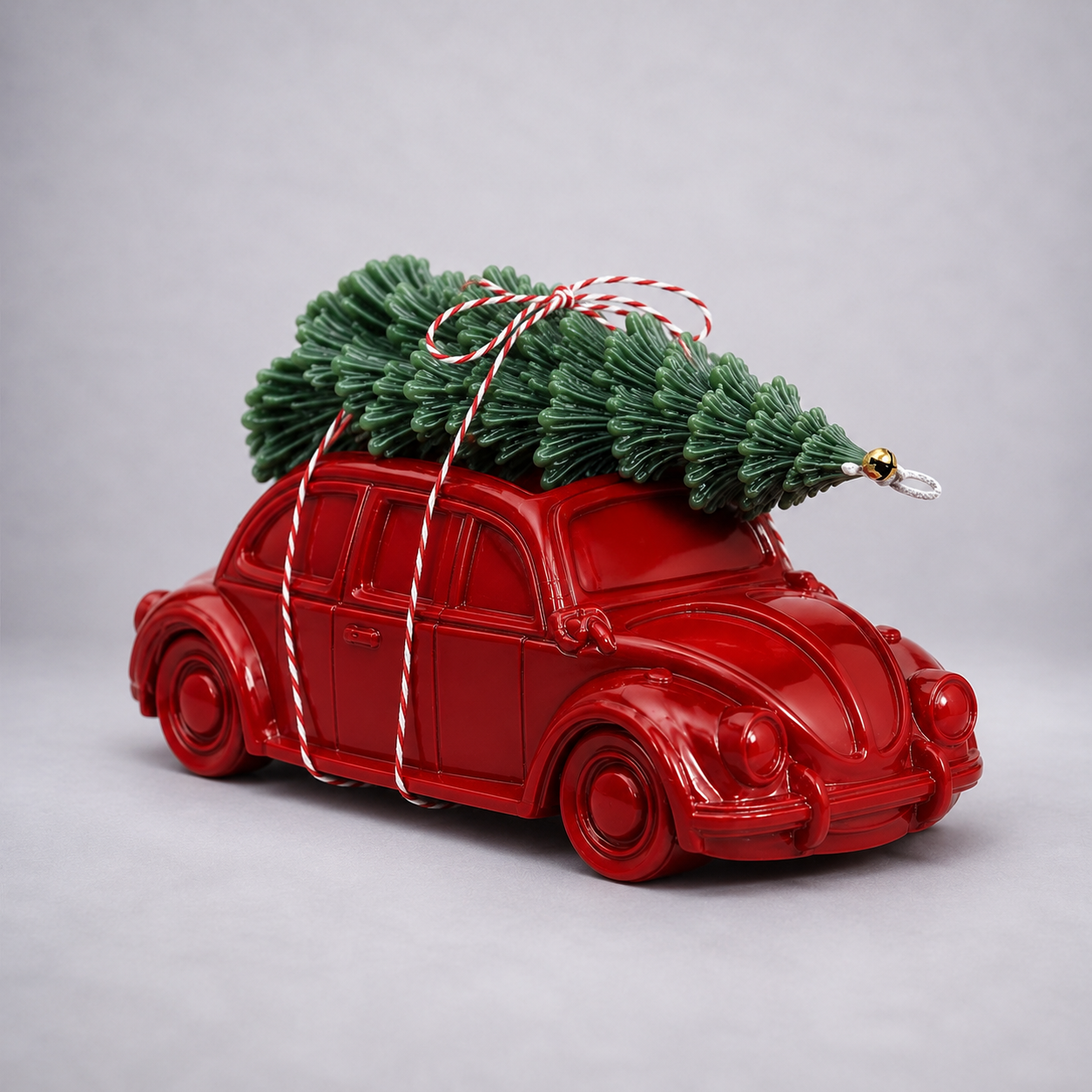 A glossy red toy Volkswagen Beetle car with a small green Christmas tree strapped on top, tied with red-and-white striped string, photographed on a light gray studio background. 
