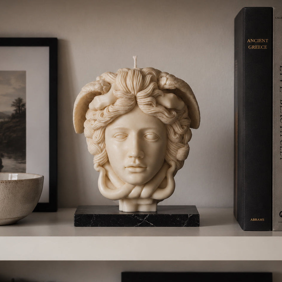 WARM WARREN HANDMADE soy wax  candle sculpted as a classical Medusa head with snakes in her hair, displayed on a black marble base on a white shelf next to framed art and books.