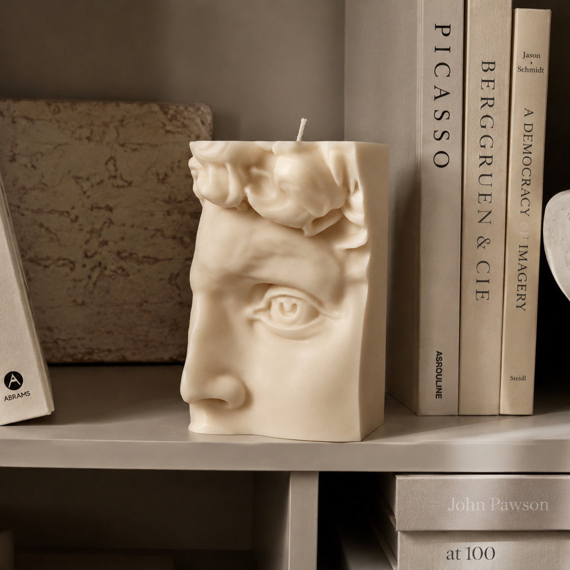 A sculptural cream-colored candle shaped like a classical male face is displayed on a bookshelf between books.