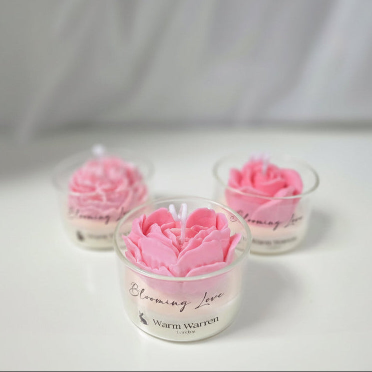 Three pink rose-shaped candles in glass containers with 'Blooming Love' branding on a white background.