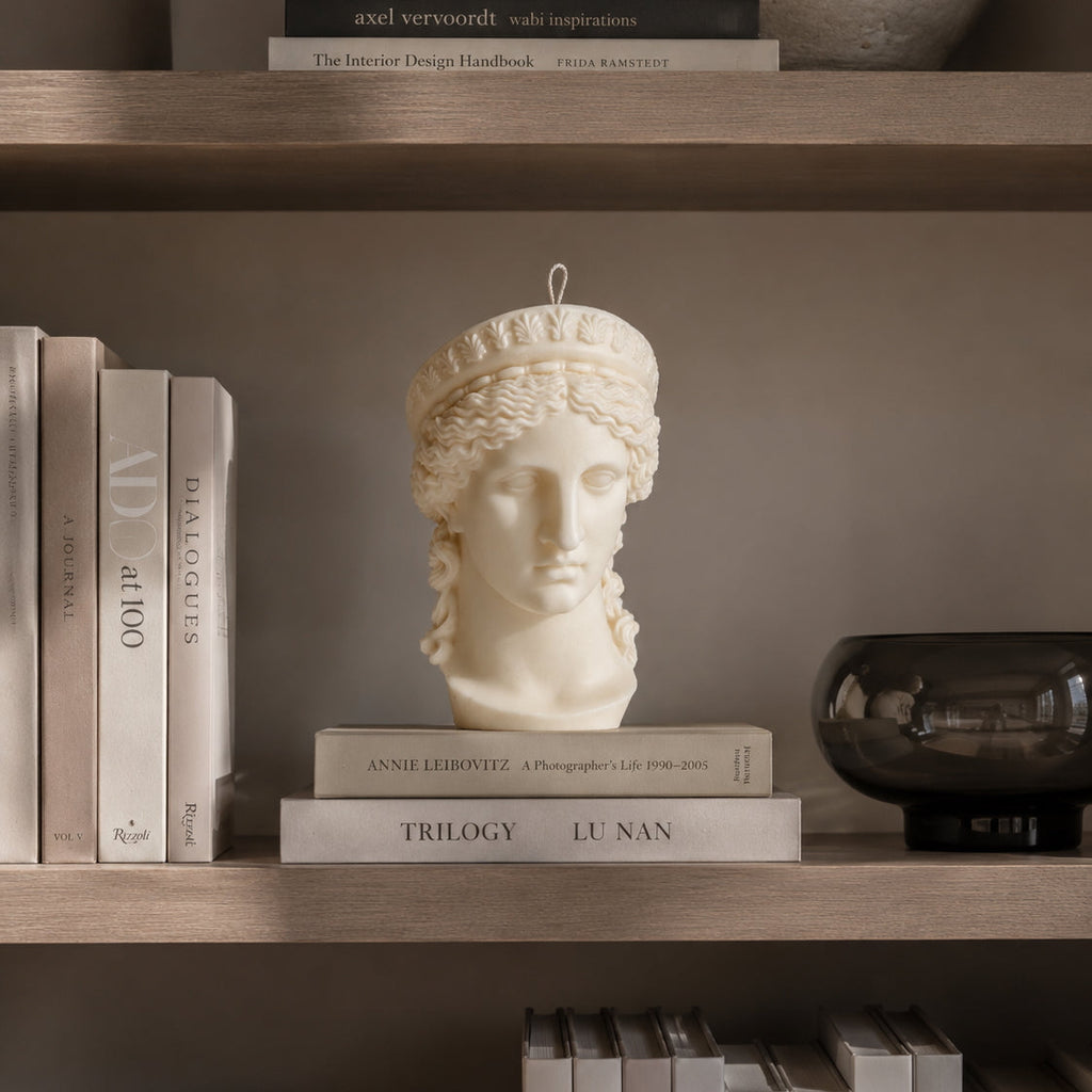 A decorative white classical goddess hera bust candle displayed on a shelf among books and home decor. Warm Warren London handmade soy wax