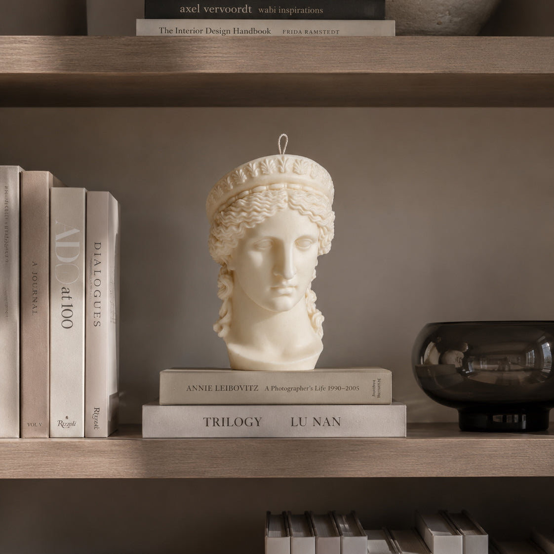 A decorative white classical goddess hera bust candle displayed on a shelf among books and home decor. Warm Warren London handmade soy wax